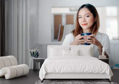 Portrait of young businesswoman holding cup and looking at camera while sitting at office desk in modern office. Wall mural