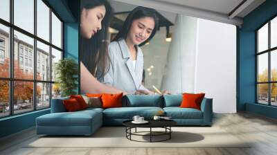 Two young asian woman discussing and working on document together while standing at office desk. Wall mural