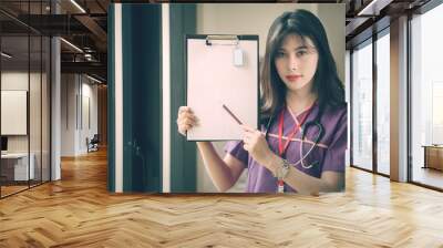 Young female nurse holding pencil and showing her clipboard Wall mural