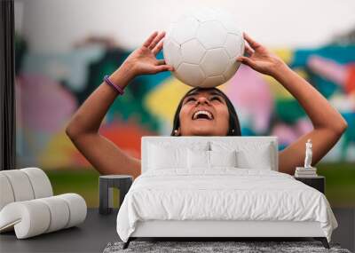 View of a beautiful brunette female soccer player posing, holding and fooling around with a soccer ball. Standing on a green grass outdoors, with a colorful background. Sportswear. Sports and wellness Wall mural