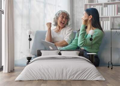 60s asian mother elderly sitting on sofa with young asia female daughter together in living room. watch movies series online or shopping, using tablet computer Wall mural