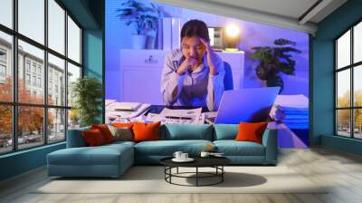 A young woman works overtime at night at her desk in office, using a laptop reviewing documents. is stressed, tired,experiencing headaches,body aches from sitting too long,leading to office syndrome Wall mural