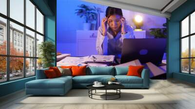 A young woman works overtime at night at her desk in office, using a laptop reviewing documents. is stressed, tired,experiencing headaches,body aches from sitting too long,leading to office syndrome Wall mural