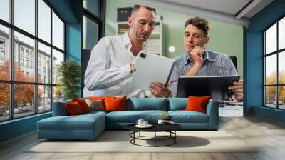 Caucasian middle-aged male businessperson and an Italian accountant are seated at a desk, engaging in a professional meeting, discussing various aspects of technology and innovation in their fields. Wall mural