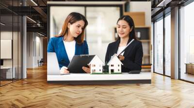 Two Asian real estate agents in formal suits discuss a contract with house models on the table, ready to sign a deal. Wall mural