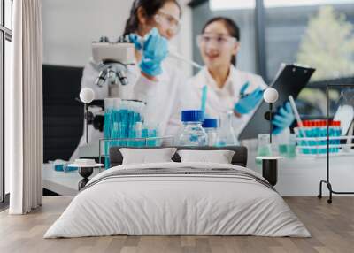 Two female researchers work at a desk in a lab, analyzing liquid samples under a microscope, experimenting with chemistry, and using high technology for important scientific discoveries. Wall mural