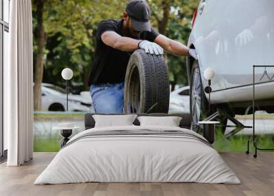 A man stands next to a car on the side of the road to fix a flat tire. He works hard to ensure safety while dealing with the damaged wheel. Wall mural