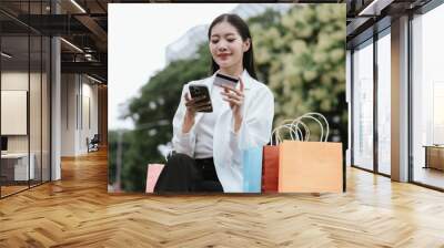 A stylish young Asian woman, cheerful and elegant, walks through a modern city mall, smiling with shopping bags. She enjoys luxury retail therapy, embodying a glamorous lifestyle. Wall mural