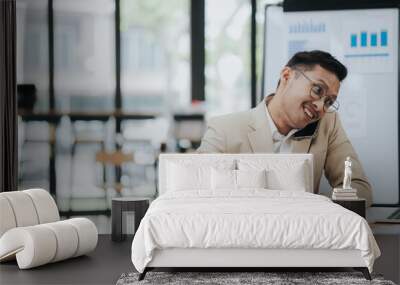 Cheerful young Asian businessman smiling in bright modern office working on laptop computer checking charts, graphs and paper. Wall mural