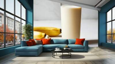 A glass of vanilla protein shake next to a banana and a scoop of protein powder on a white surface. Wall mural