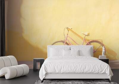 A pastel pink bicycle parked against a pale yellow wall with soft sunlight. Wall mural