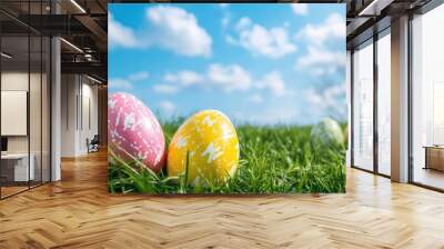 Colorful Easter eggs resting on a bed of fresh green grass, with a clear blue sky and fluffy clouds. Wall mural