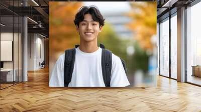 young man smiles confidently while walking outdoors, surrounded by vibrant autumn foliage. His casual attire and backpack suggest relaxed day, capturing moment of joy and youthfulness. Wall mural