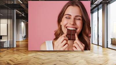 A beautiful woman in white shirt eats chocolate bar isolated on pink background, happy face expression with closed eyes and mouth open , stock photo, studio light Wall mural