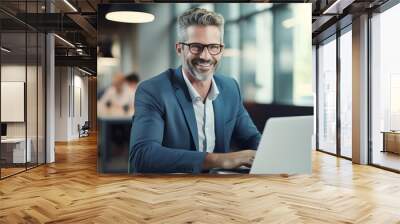 A smiling adult businessman who is using a laptop while seated at his desk. A contented and active middle-aged businessman, CEO, and manager who works in an office using corporate technology Wall mural