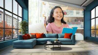 leisure, education and home concept - smiling teenage girl reading book and sitting on couch at home. Portrait of a young woman lying on couch with book. A teenage girl with a book in library. Wall mural