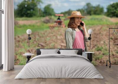 Young woman standing on field in sunset while tractor baling in background, Farmer or agrarian with smart phone looking on growing efficiency and crop yield, Smart agriculture and automation concept. Wall mural