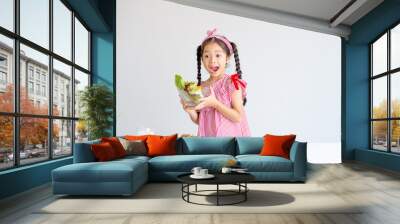 Asian little cute girl show action of surprise and hold bowl of vegetables with the apples and bread on table. Wall mural