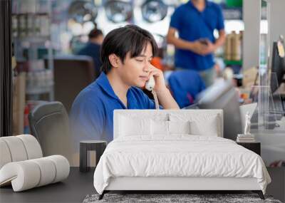 Asian man receive phone call from customer and stay in counter service area of automotive or garage shop with other worker work in the background. Wall mural