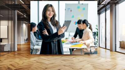 Portrait of young Asian business woman hold tablet and stand with look at camera in front of other co-workers discuss on the back in office. Wall mural