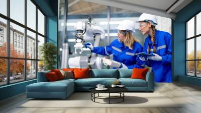 Two Caucasian professional engineer or technician woman workers hold controller and tablet help to check and maintenance robotic arm machine with stand beside in factory workplace. Wall mural