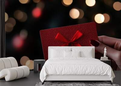 A beautifully wrapped red gift card with a bow is held against a dreamy bokeh background of shimmering holiday lights, embodying the joy of giving and festive cheer. Wall mural