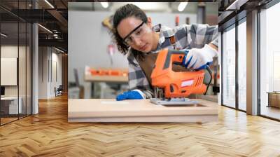 Beautiful Latin woman carpenter using power tools working her wood job in carpenter's shop. Young hispanic female in protective goggles busy in furniture woodworking. Feminism in carpentry industry. Wall mural