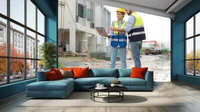 Engineer man and female architect wear safety helmets discuss housing development project at construction site using laptop computer. Contractor manager examining building estate infrastructure. Wall mural