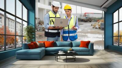 Engineer man and female architect wear safety helmets discuss housing development project at construction site using laptop computer. Contractor manager examining building estate infrastructure. Wall mural