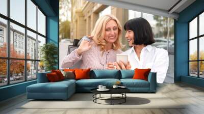 Two mature women looking at the phone and having fun while doing shopping Wall mural