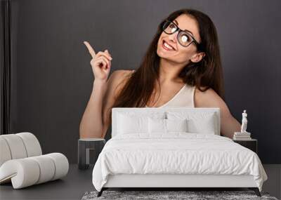 Beautiful positive business happy woman in eyewear in white t- shirt showing up and  demonstrate idea sign by fingers above on empty copy space grey background. Closeup Wall mural