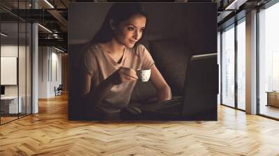 Smiling thinking beautiful woman looking on computer monitor drinking coffee on dark shadow home background in the evening. Wall mural