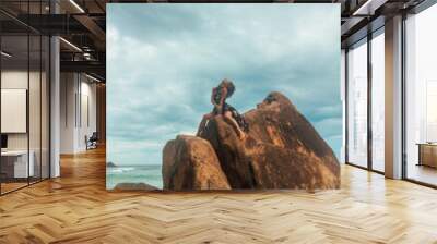 holiday leisure. young brunette girl in black stylish dress is sitting fashion with flirt and looking up on a huge rock stone on blue sky background with ocean. travel lifestyle concept, free space Wall mural