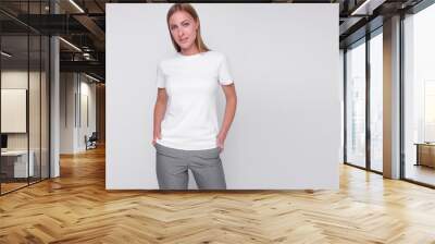 young stylish blonde girl in gray pants and white t-shirt is standing with hands in her pockets and smiling cute on the white wall background. medical concept. free space on right side, mockup Wall mural