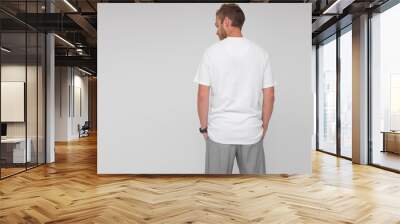 young stylish male doctor in white t-shirt and gray pants is standing from the back with hands in pockets on the white wall background. medical concept. free space on left side, mockup Wall mural