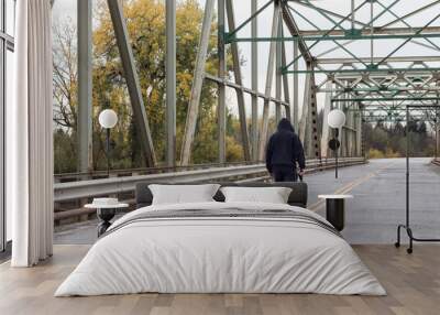 horizontal image of a very troubled man walking in the middle of the road over a steel bridge carrying a small pistol  on a grey cool fall day. Wall mural