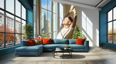 Enjoying new day. Beautiful happy mature woman stretching body and smiling while standing with closed eyes in sun rays shining through window on sunny morning at home. Optimism and happiness concept Wall mural