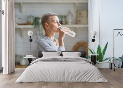 Side view of beautiful senior woman drinking yogurt and looking through the window while standing in the modern kitchen at home. Mature people and healthy eating concept Wall mural