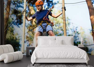 Child in forest adventure park. Kid in orange helmet  and blue t shirt climbs on high rope trail. Agility skills and climbing outdoor amusement center for children. young boy plays outdoors. Wall mural