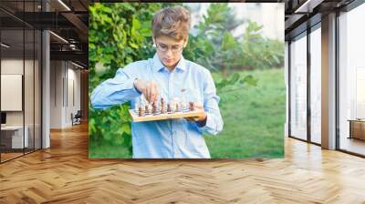 cute young boy in blue shirt holds chess board  and plays chess in the summer park. Education, intellectual game  Wall mural