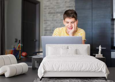 Cute young teenager in yellow shirt sitting behind desk in kitchen next to laptop and study. Serious boy makes homework, listening lesson. Home, distance education, self study by kids. Wall mural