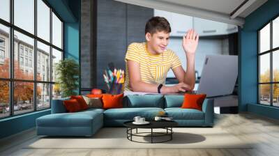 Cute young teenager in yellow shirt sitting behind desk in kitchen next to laptop and study. Serious boy makes homework, listening lesson. Home, distance education, self study by kids. Wall mural