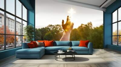 A hand is reaching a glowing cross image which is situated in the sky above the green summer trees. Wall mural