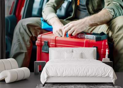 A person sitting in a vehicle while handling a red first aid kit with a white cross, likely preparing for an emergency situation or medical assistance. Wall mural