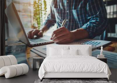 A professional working on a laptop with documents and charts on a wooden desk, highlighting analysis, data review, and business planning in a sunlit office. Wall mural