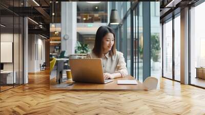 Asian woman working on laptop in a modern office with bright natural light, stylish furniture and large windows. Casual business environment. Wall mural