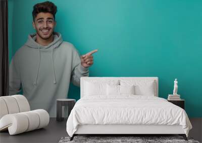 Cheerful young man in a gray hoodie pointing sideways on a turquoise background. Wall mural