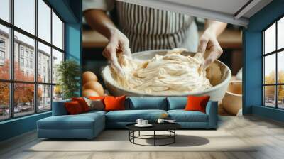 Close-up of a person mixing dough in a large bowl with hands, surrounded by ingredients like eggs and kitchen utensils on a wooden countertop. Wall mural