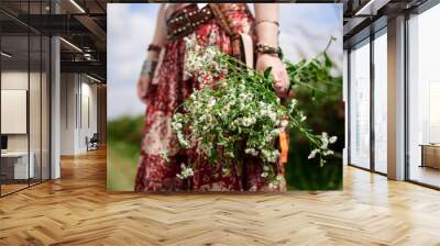 Close-up picture of wildflowers in female hands. Young hippie woman, wearing colorful boho style clothes, standing on green field, holding camomiles' bouquet. Eco tourism concept. Wall mural