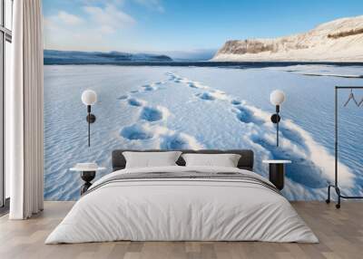 Footprints in deep snow on a vast frozen lake with a backdrop of snowy mountains and a clear blue sky. Wall mural